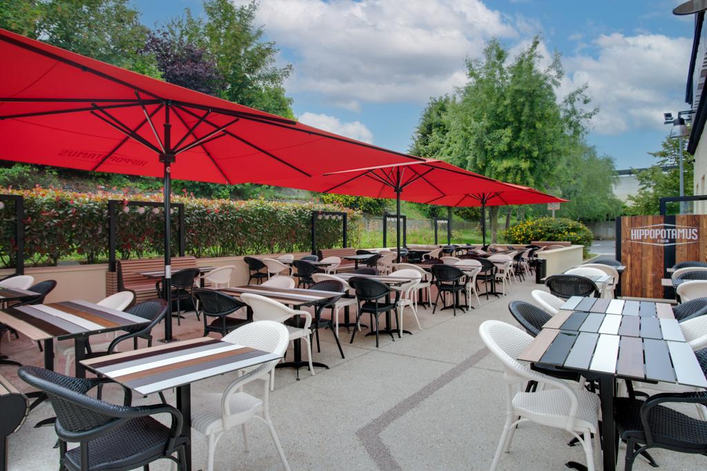 Terrasse extérieure d'un restaurant Hippopotamus avec des rangées de tables et de chaises sous des parasols rouge vif, entourée de verdure et d'un ciel bleu nuageux.