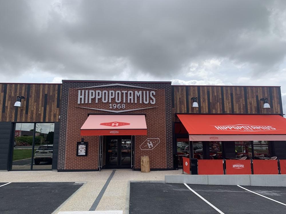 Extérieur du restaurant avec l'enseigne Hippopotamus, avec des auvents rouges, une façade en brique et en bois, et un ciel nuageux au-dessus.