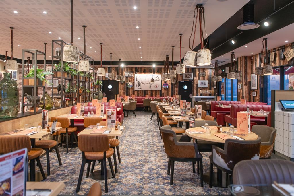 Un restaurant Hippopotamus moderne avec des tables en bois, des chaises en cuir, des suspensions lumineuses et des éléments décoratifs, créant une atmosphère chaleureuse et accueillante.