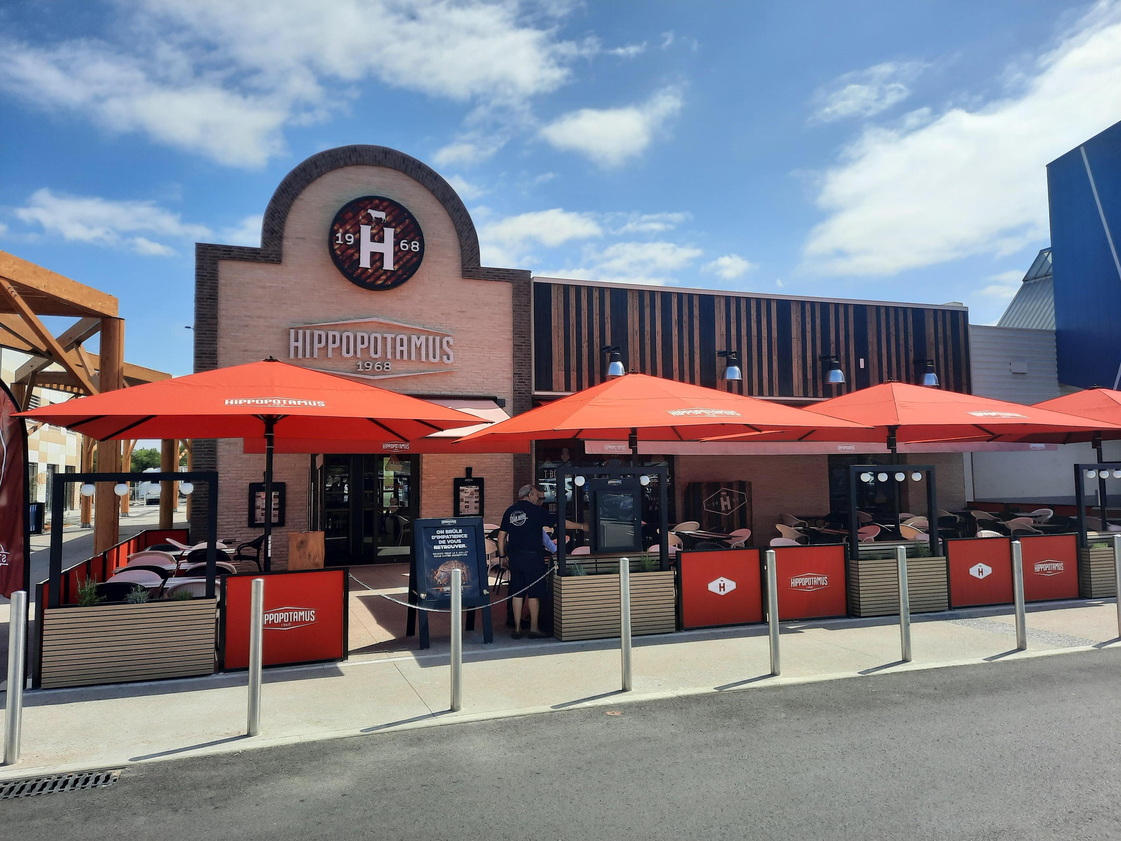 Extérieur d'un restaurant Hippopotamus avec une façade en briques et des parasols rouges. Ciel bleu en arrière-plan.