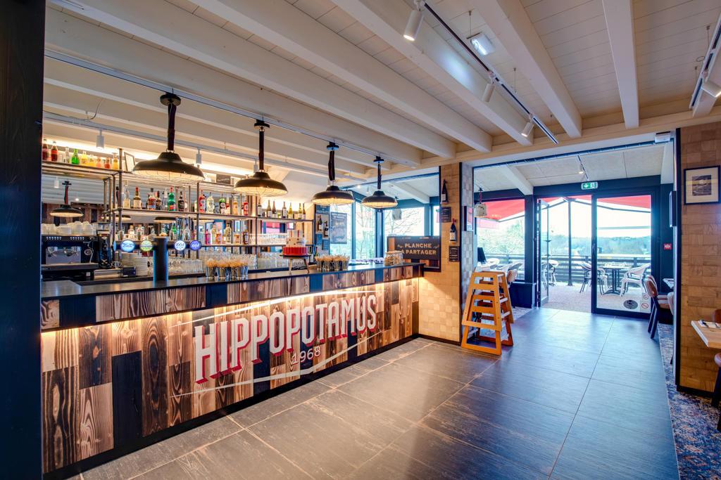 Intérieur d'un restaurant avec un restaurant Hippopotamus, avec des étagères remplies de bouteilles, un éclairage industriel et une entrée ouverte sur une terrasse.