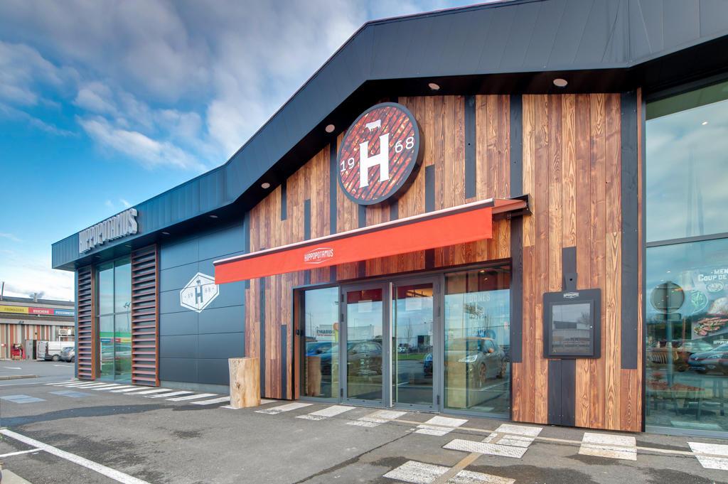 Extérieur moderne d'un restaurant Hippopotamus avec des panneaux en bois et noirs, un auvent rouge et un ciel clair en arrière-plan.