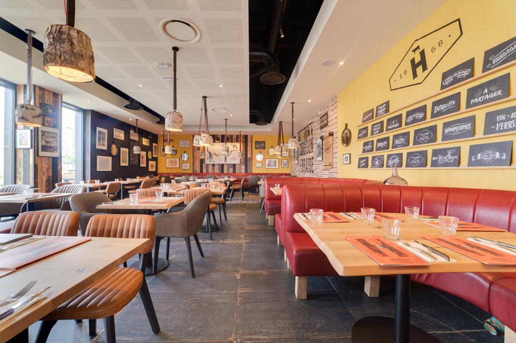 Intérieur lumineux du restaurant Hippopotamus avec des banquettes rouges, des tables en bois dressées avec des couverts, des murs jaunes ornés de divers tableaux encadrés et décorations.