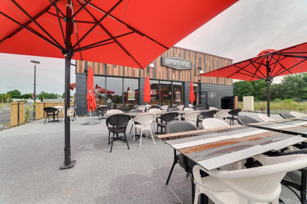 Terrasse d'un restaurant Hippopotamus avec des parasols rouges, des chaises noires et blanches et des tables en bois sous un ciel nuageux.