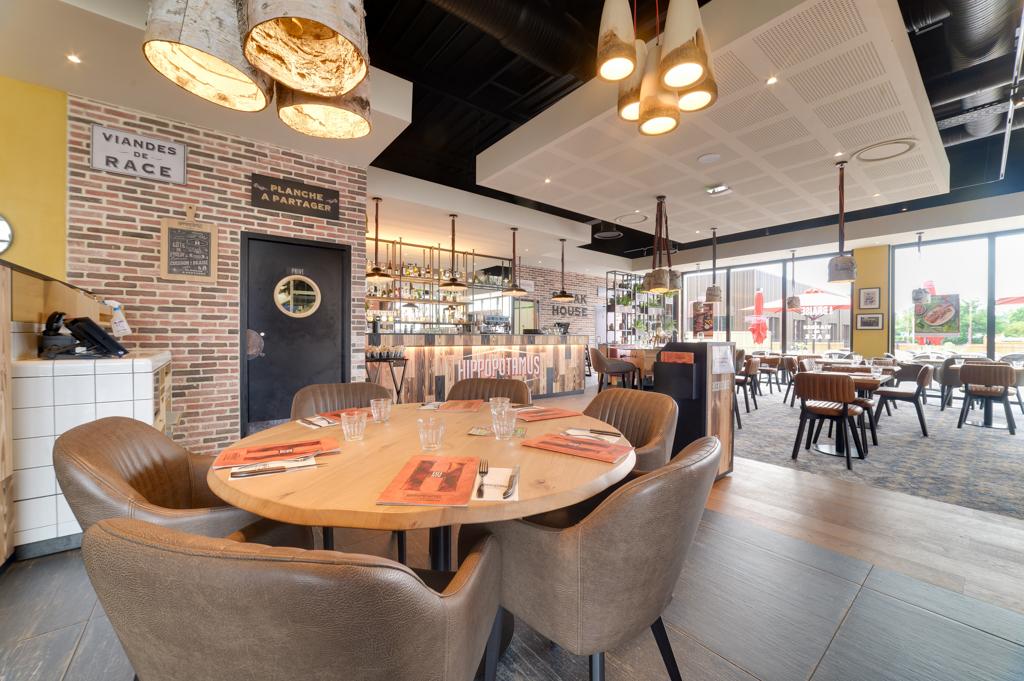 Intérieur moderne d'un restaurant Hippopotamus avec des murs en briques, une table ronde en bois dressée pour le dîner, des chaises en cuir, des suspensions lumineuses et un bar à l'arrière-plan.