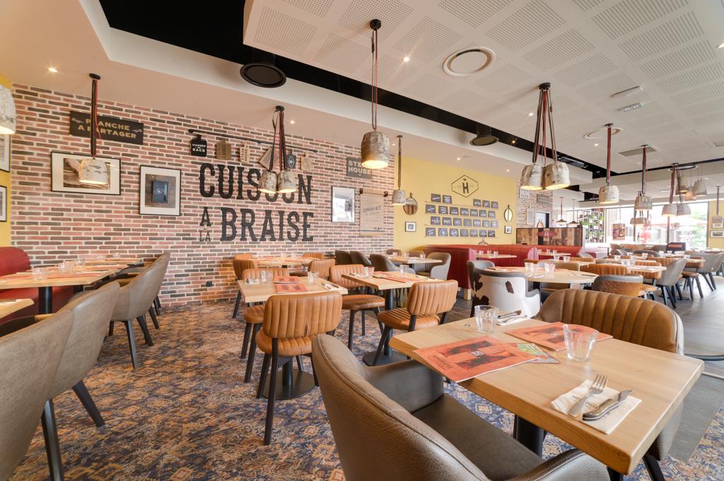 Restaurant Hippopotamus à l'intérieur chaleureux avec des tables en bois, des chaises rembourrées, un mur en briques apparentes, des suspensions lumineuses et des tables dressées pour accueillir les clients.
