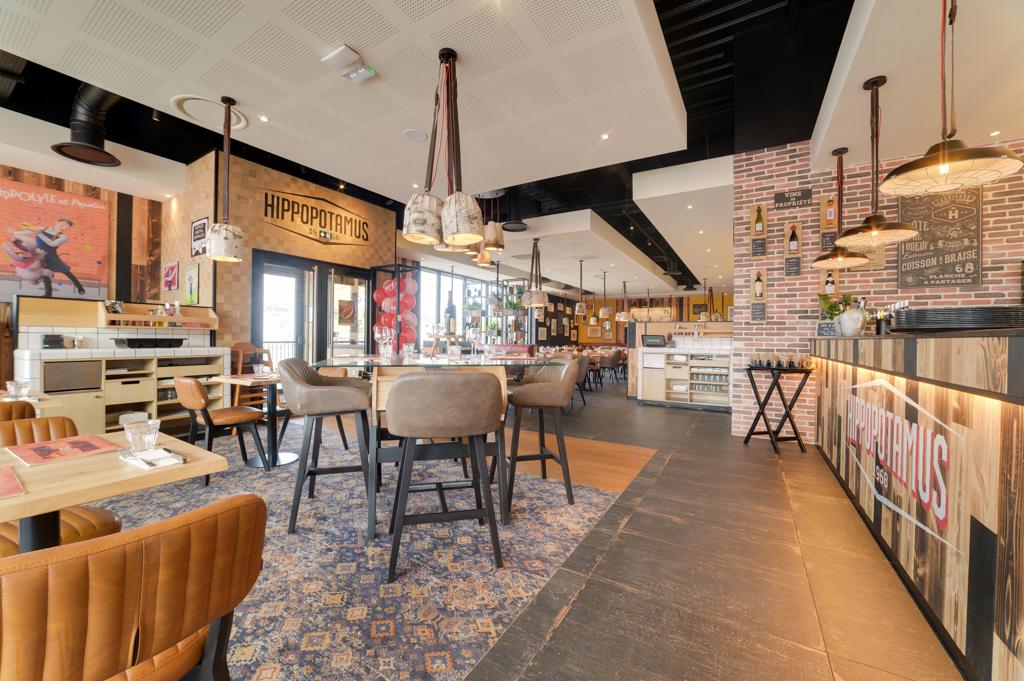 Restaurant Hippopotamus spacieux à la décoration moderne, avec des tables en bois, des chaises rembourrées, des suspensions lumineuses et un tapis à motifs, le tout dans une ambiance chaleureuse.