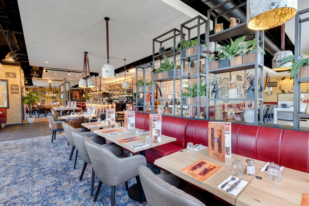 Intérieur spacieux du restaurant avec des banquettes rouges, des tables en bois sur lesquelles sont disposés les menus et des plantes décoratives sur des étagères. Ambiance chaleureuse et moderne.
