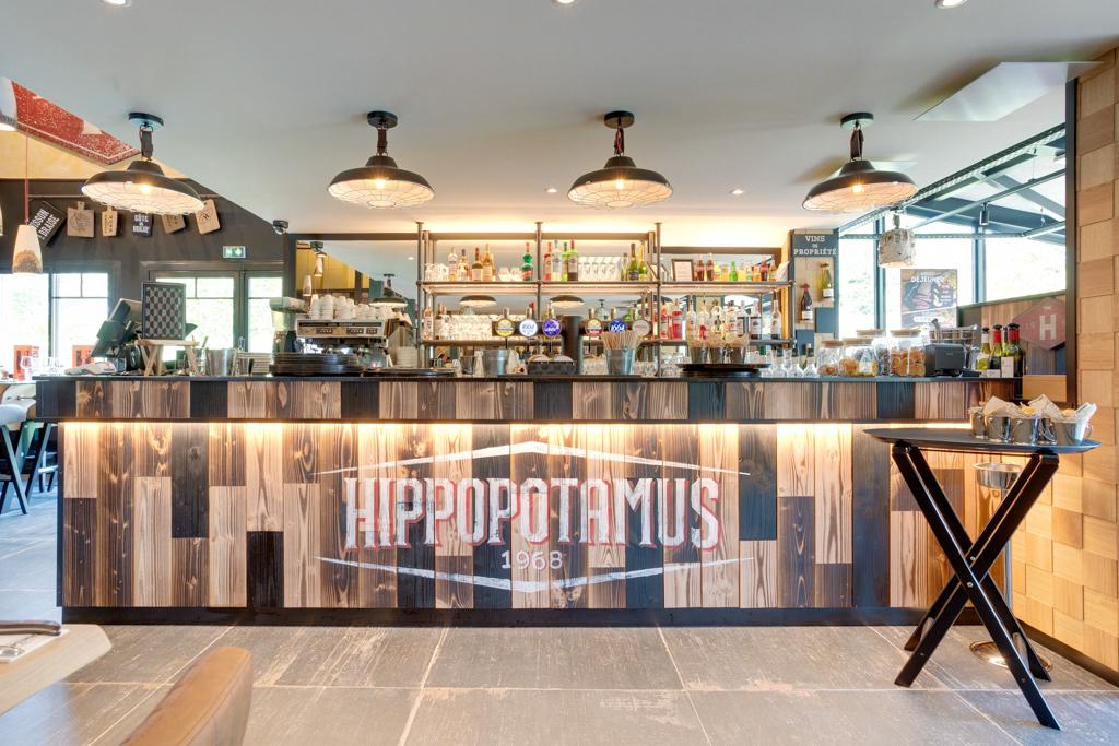 Restaurant Hippopotamus moderne, avec un bar à panneaux en bois rustiques, plafonniers et étagères remplies de bouteilles et de verres.