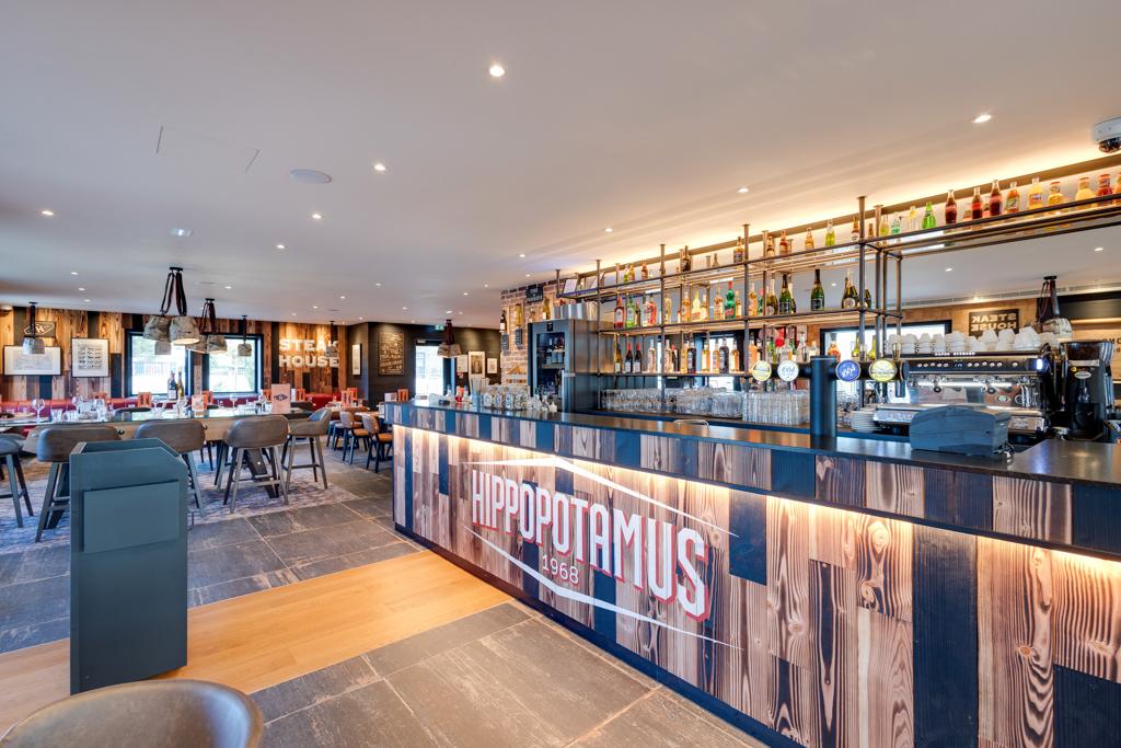 Intérieur moderne d'un restaurant Hippopotamus avec comptoir en bois, tabourets de bar, étagères remplies de bouteilles et éclairage chaleureux