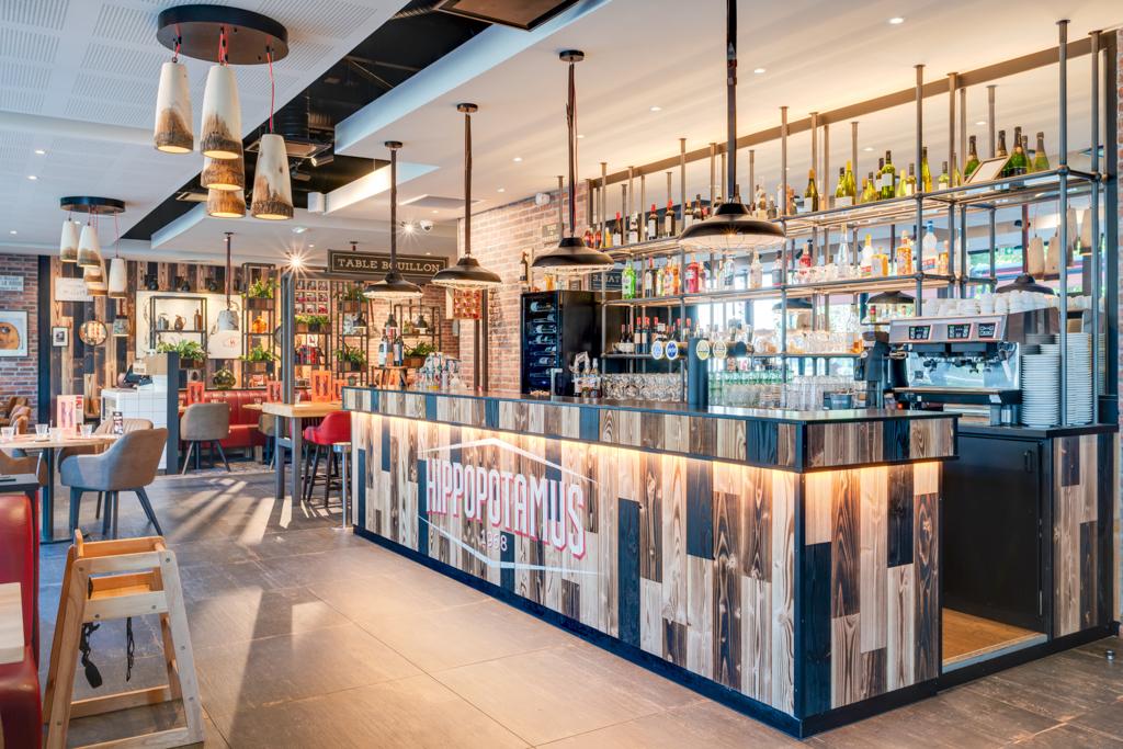 Intérieur moderne d'un restaurant Hippopotamus avec un bar coloré, des suspensions lumineuses et des étagères remplies de bouteilles. Accents en bois et tables avec chaises.