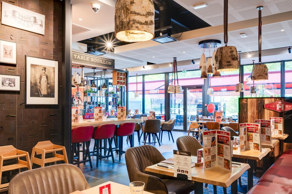 Restaurant Hippopotamus à l'intérieur chaleureux avec des tables en bois, des chaises rouges et beiges, des suspensions lumineuses et de grandes fenêtres laissant entrer la lumière naturelle.