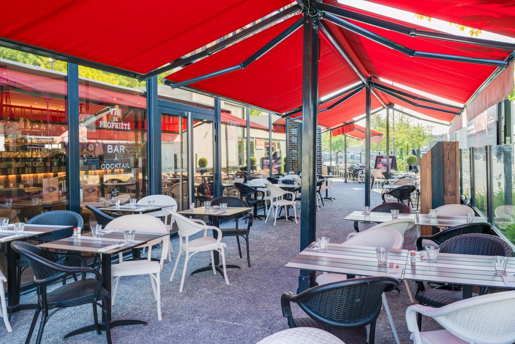 Restaurant Hippopotamus en plein air avec des auvents rouges, des chaises noires et blanches, des tables en verre et des tables dressées pour le déjeuner, entouré de verdure et de baies vitrées.