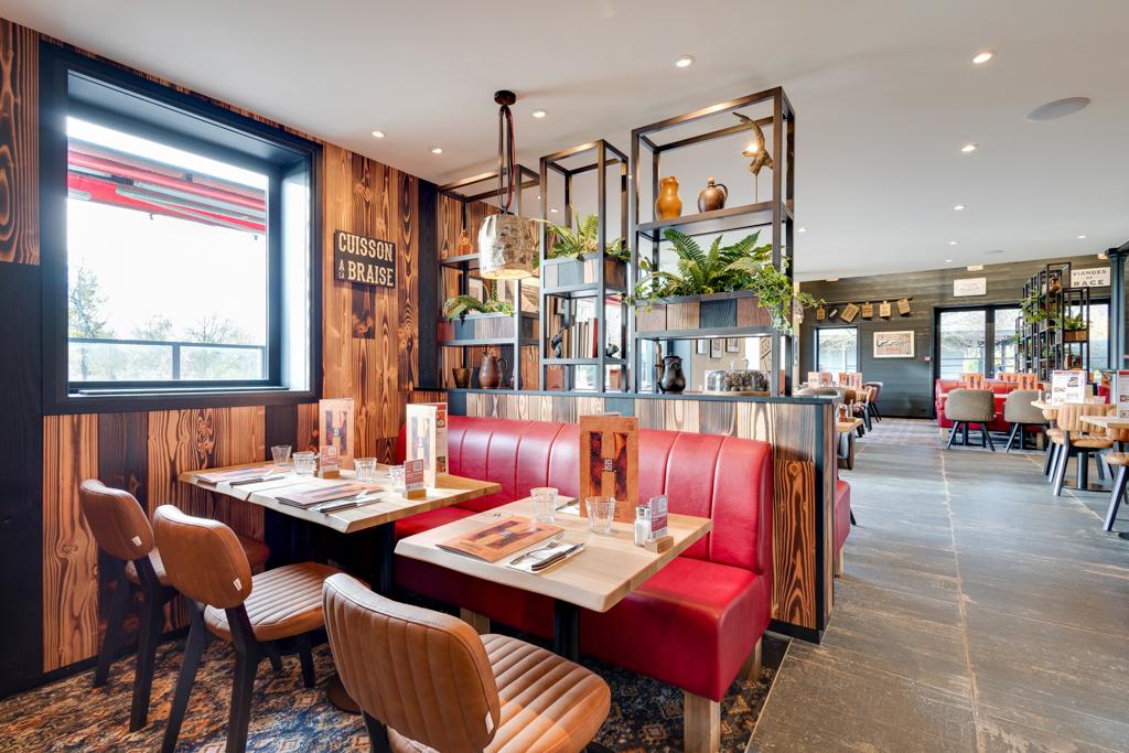 Intérieur chaleureux avec banquettes rouges, murs en bois, plantes décoratives et tables dressées pour le dîner. De grandes fenêtres laissent entrer la lumière naturelle.