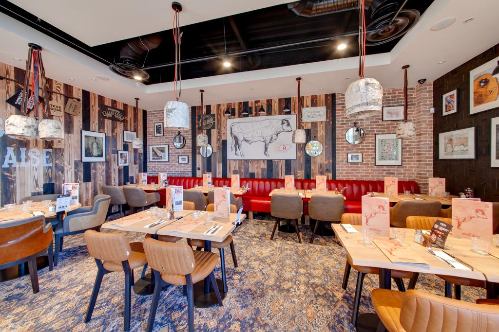 Intérieur chaleureux d'un restaurant Hippopotamus avec murs en bois, sièges en cuir rouge, suspensions modernes et tables dressées pour le dîner. Œuvres d'art et décorations représentant des vaches aux murs.