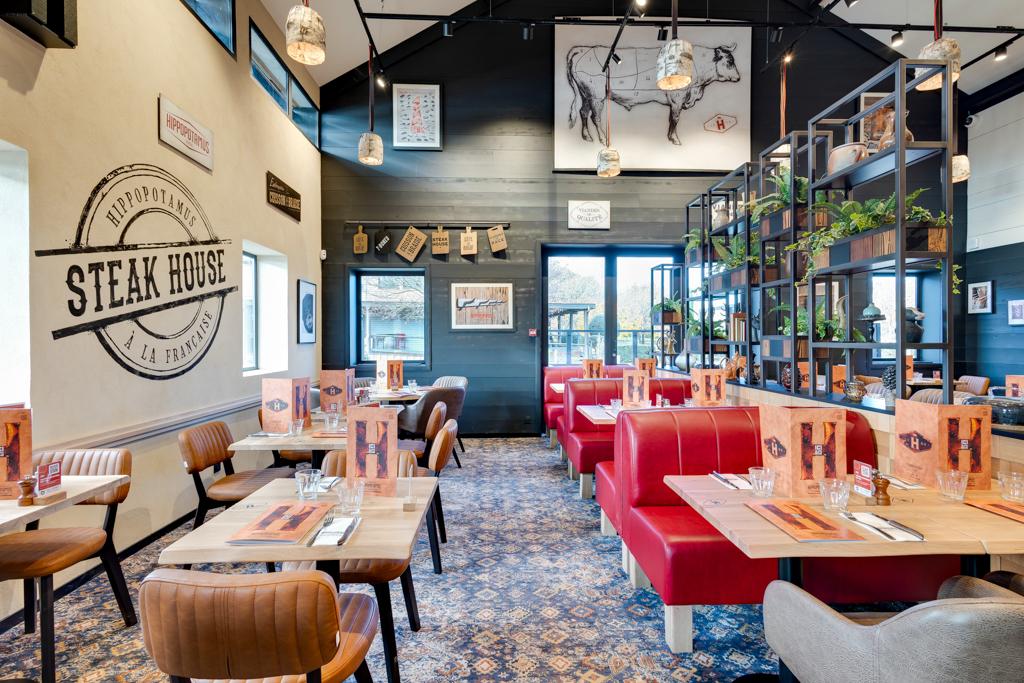 Intérieur moderne de steakhouse avec banquettes rouges, tables en bois et décorations murales. Les plantes suspendues et les suspensions lumineuses créent une atmosphère chaleureuse.