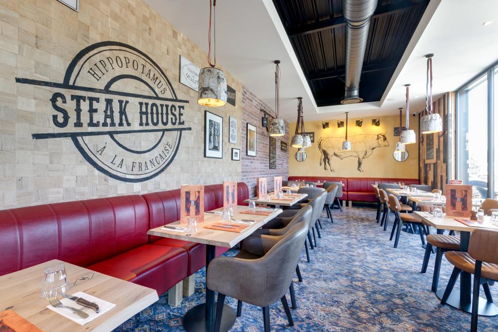 Intérieur lumineux du restaurant Hippopotamus avec banquettes rouges, tables en bois et suspensions lumineuses. Les décorations murales et les grandes fenêtres renforcent l'atmosphère chaleureuse.