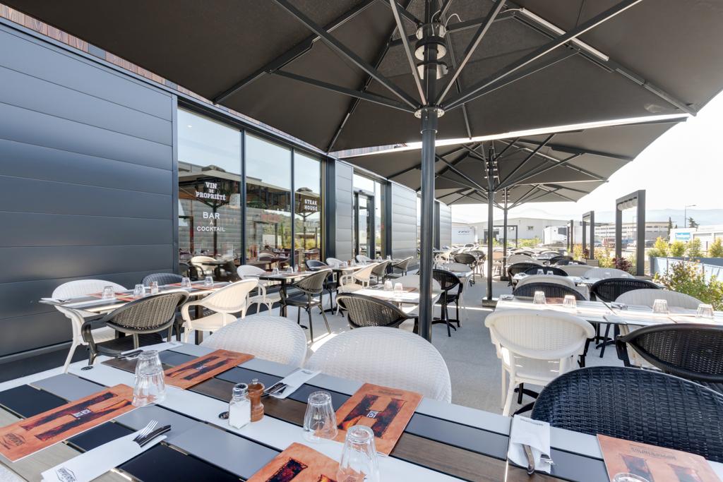 Terrasse de restaurant Hippopotamus à l'extérieur avec tables dressées pour le dîner, chaises noires et blanches, grands parasols et bâtiment moderne en arrière-plan.