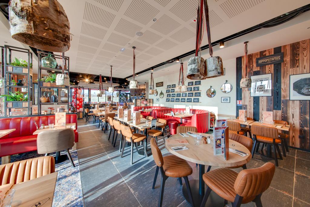 Intérieur moderne de restaurant Hippopotamus Steakhouse avec des sièges rouges et beiges, des tables en bois et des suspensions décoratives. Murs ornés de photos et de plantes.