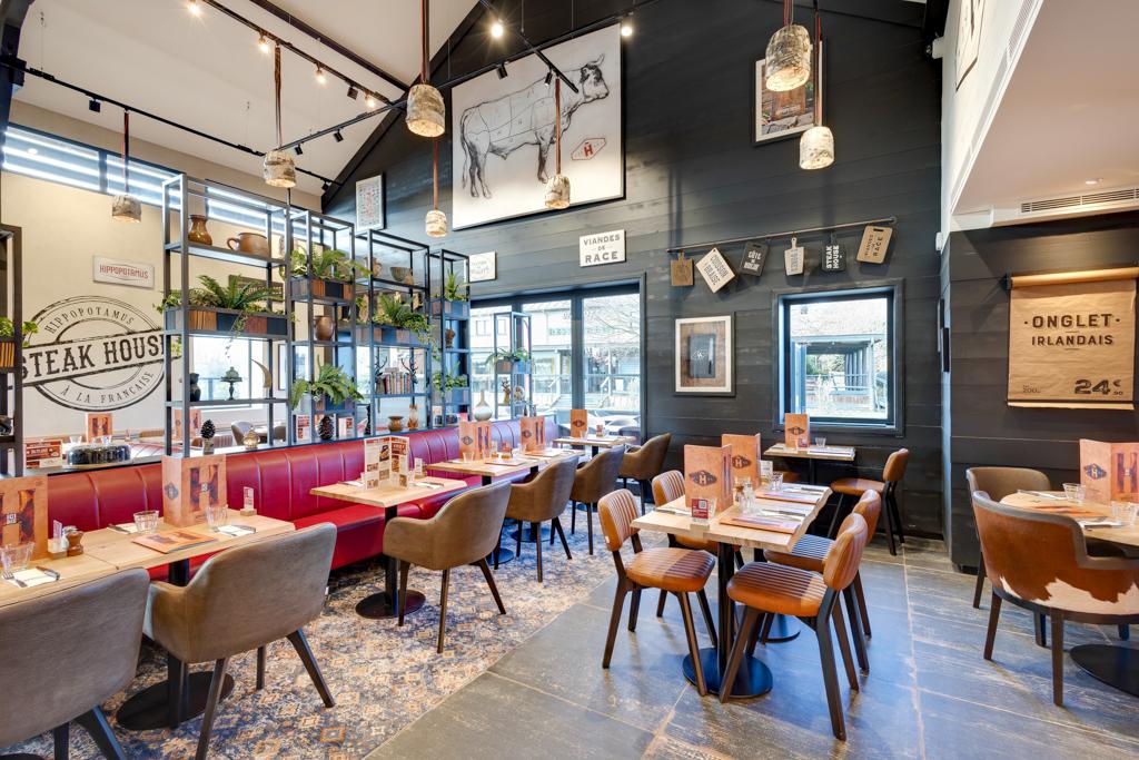 Intérieur chaleureux du restaurant Hippopotamus avec tables en bois, chaises moelleuses, décorations murales, suspensions lumineuses et étagère décorée de plantes.