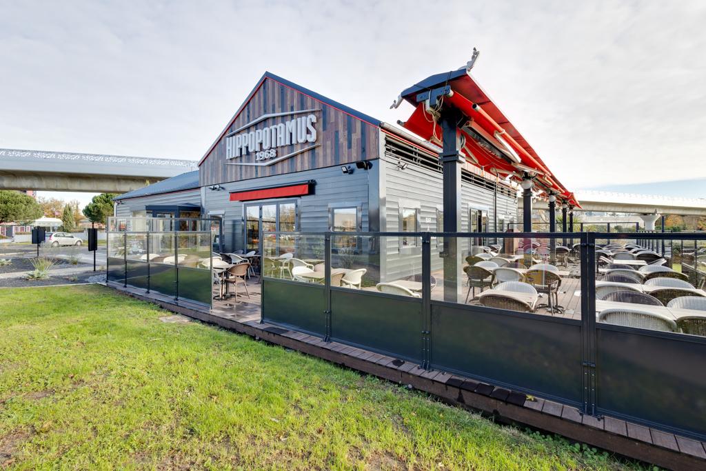 Un restaurant Hippopotamus moderne avec une façade rouge et grise, des tables en terrasse sous un auvent rouge et une clôture en verre entourant le patio.