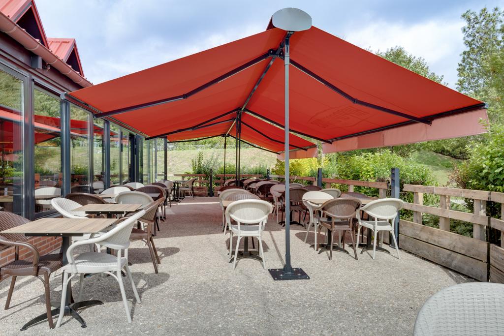 Terrasse extérieure avec auvent rouge, tables et chaises vides, entourée de baies vitrées et de verdure par une journée ensoleillée.