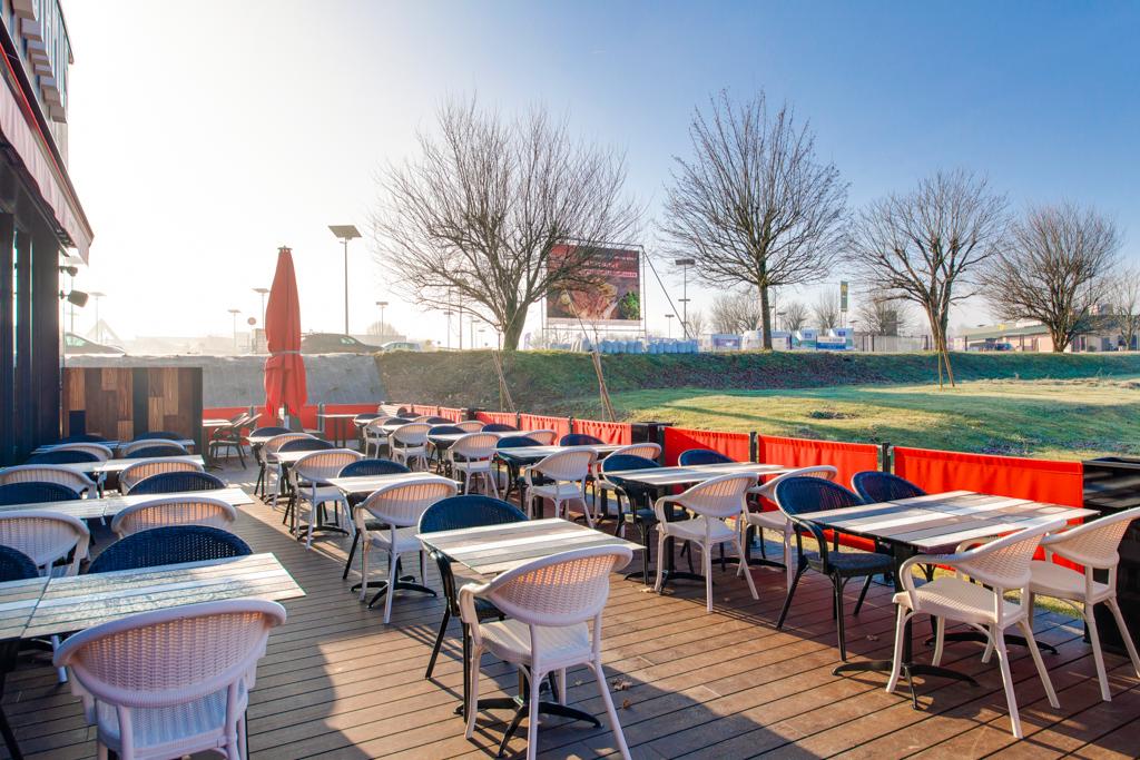 Terrasse de restaurant Hippopotamus en plein air avec tables et chaises vides, parasols rouges et pelouse avec des arbres dénudés sous un ciel bleu clair.