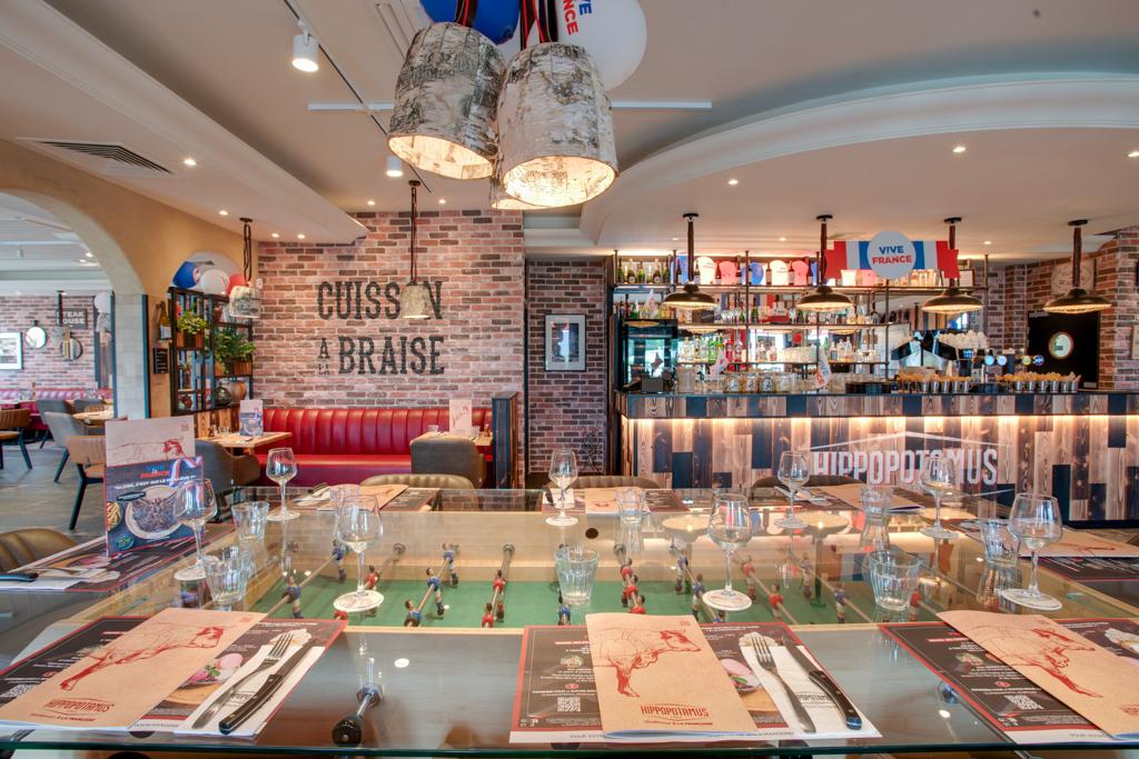 Restaurant Hippopotamus confortable avec une table de baby-foot servant de table à manger, des murs en briques, des banquettes rouges et un bar décoré de drapeaux et d'objets divers.