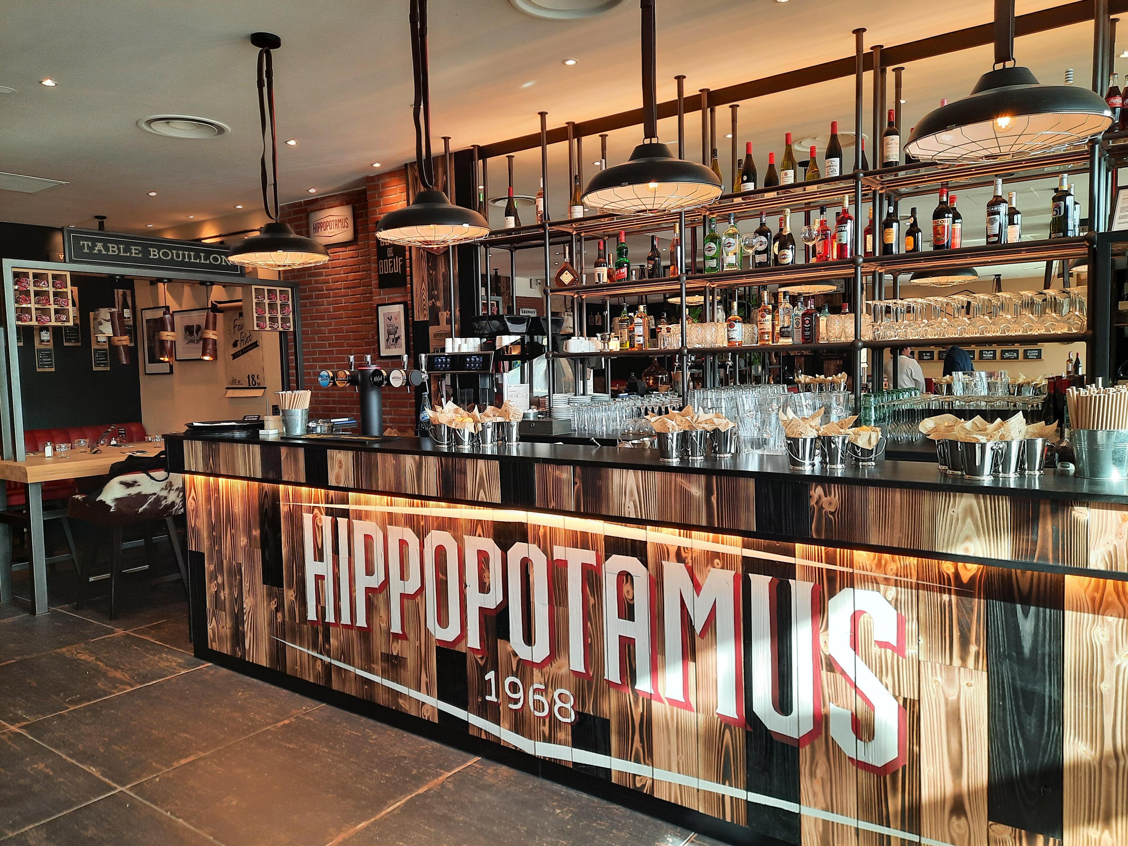 Un bar moderne avec un comptoir en bois portant l'inscription « Hippopotamus 1968 », équipé de suspensions lumineuses, de tabourets de bar et d'étagères remplies de bouteilles et de verres.