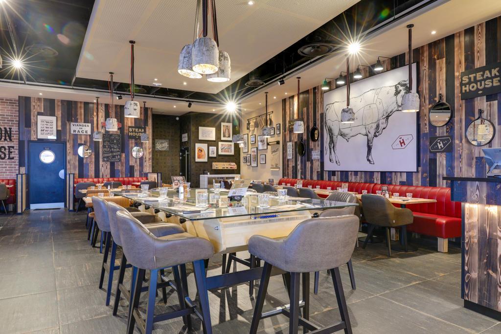 Intérieur de restaurant tendance Hippopotamus avec une décoration moderne, comprenant une longue table, des chaises rembourrées, des œuvres d'art représentant des vaches et des banquettes rouges contre des murs en bois.