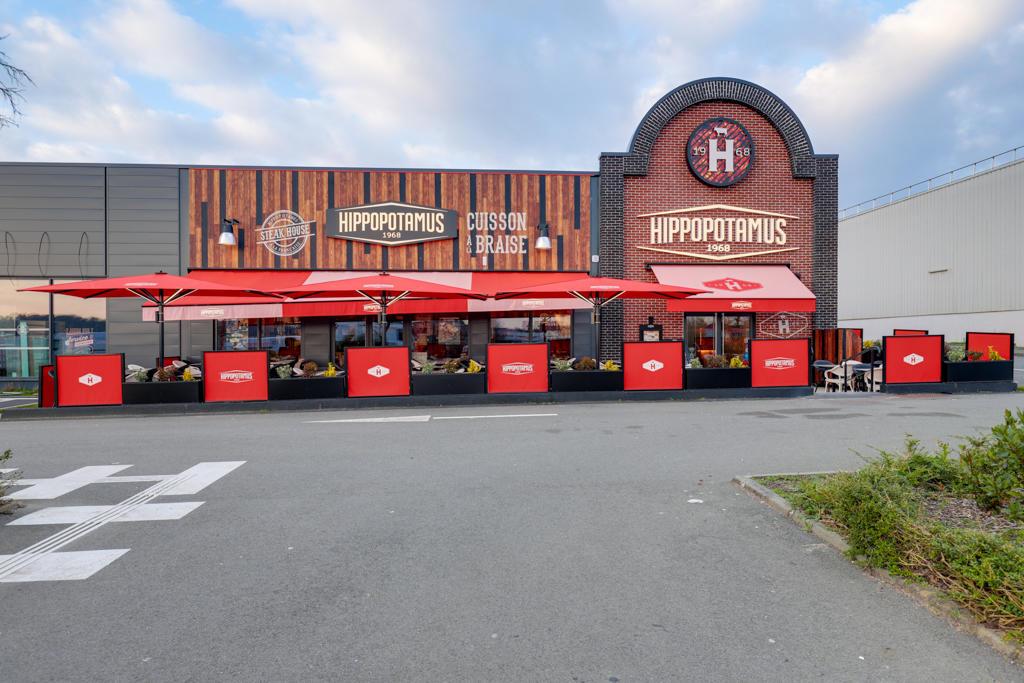 Extérieur d'un restaurant Hippopotamus avec des parasols rouges sous un ciel nuageux. Terrasse extérieure visible.