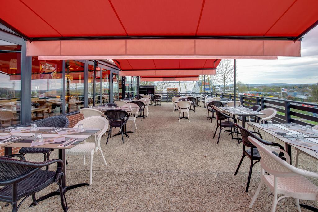 Terrasse extérieure d'un restaurant Hippopotamus avec des auvents rouges, plusieurs tables, des chaises noires et blanches, et une vue sur des arbres et un ciel nuageux en arrière-plan.