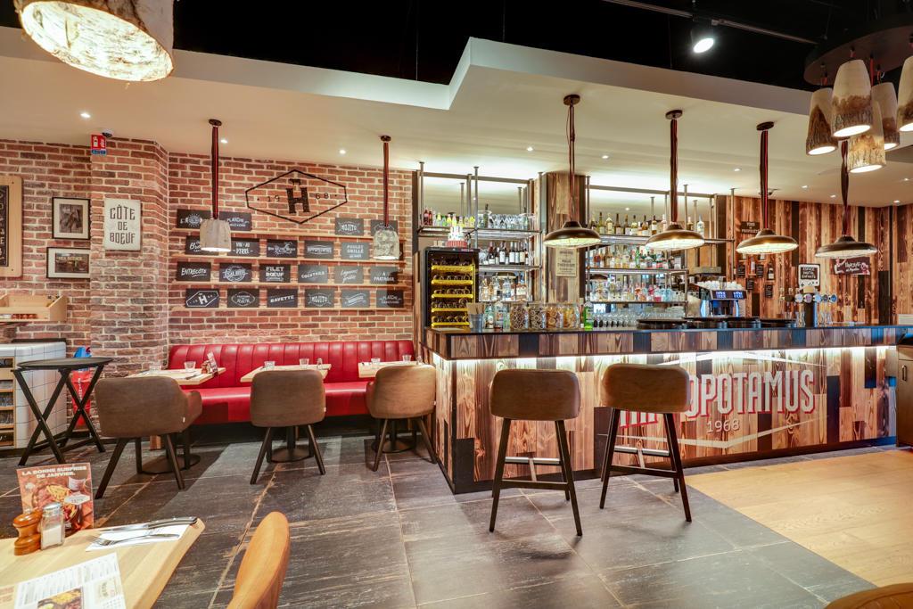 Intérieur chaleureux d'un restaurant Hippopotamus avec un mur en briques, des banquettes rouges, un bar aux accents boisés et un éclairage tamisé créant une atmosphère chaleureuse.
