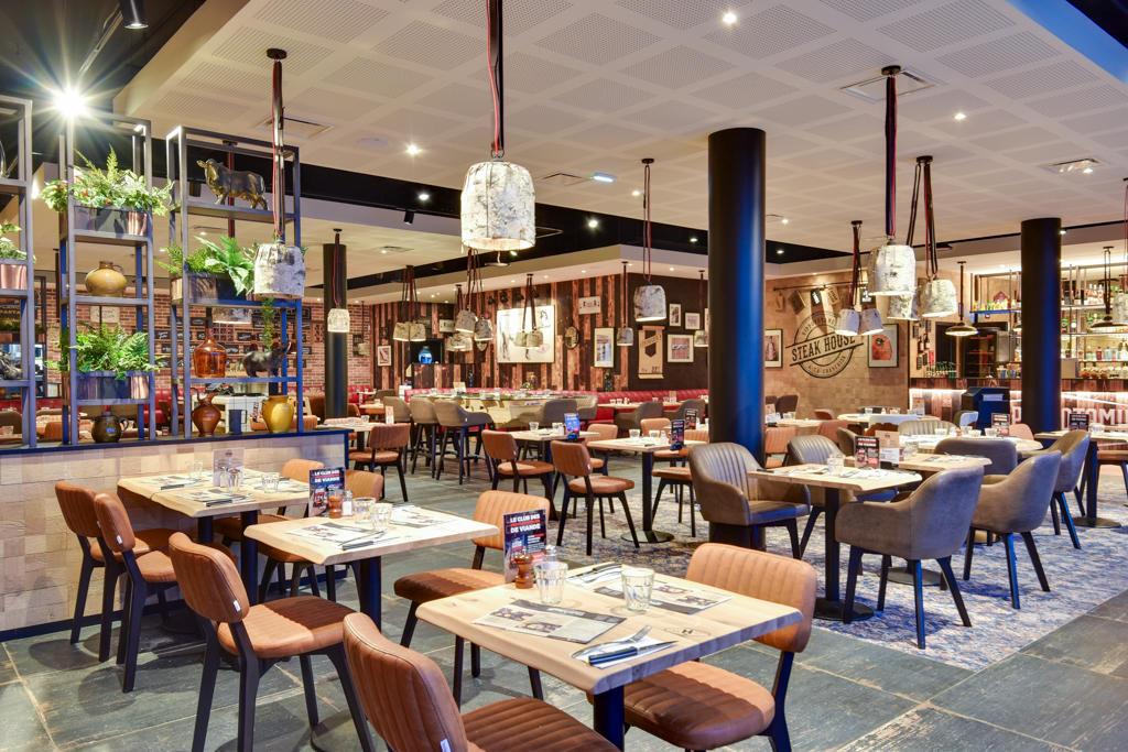 Intérieur moderne de restaurant Hippopotamus avec tables en bois, chaises rembourrées, suspensions lumineuses et plantes décoratives. Ambiance chaleureuse et accueillante.