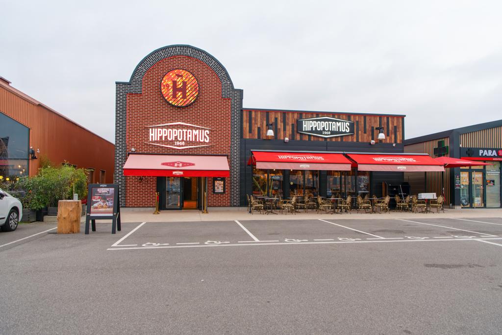 Extérieur d'un restaurant Hippopotamus avec des auvents rouges, des tables en terrasse et des places de stationnement devant, sous un ciel nuageux.