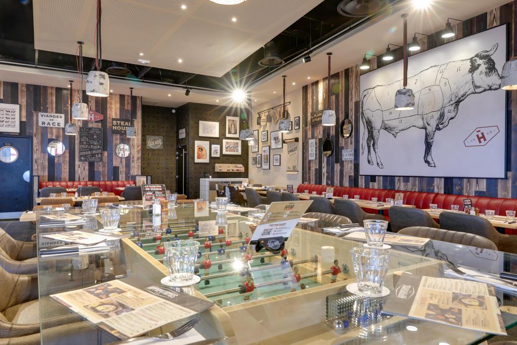 Un restaurant Hippopotamus à l'intérieur élégant avec des murs en bois, des œuvres d'art représentant des vaches, des sièges rouges et des tables dressées avec des menus et des verres. On aperçoit un baby-foot.