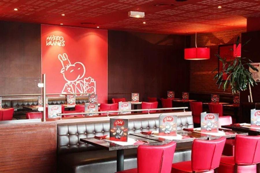 Intérieur de restaurant Hippopotamus avec une décoration rouge et marron, comprenant des banquettes rouges, des tables dressées avec des menus et un personnage sur un grand panneau mural.
