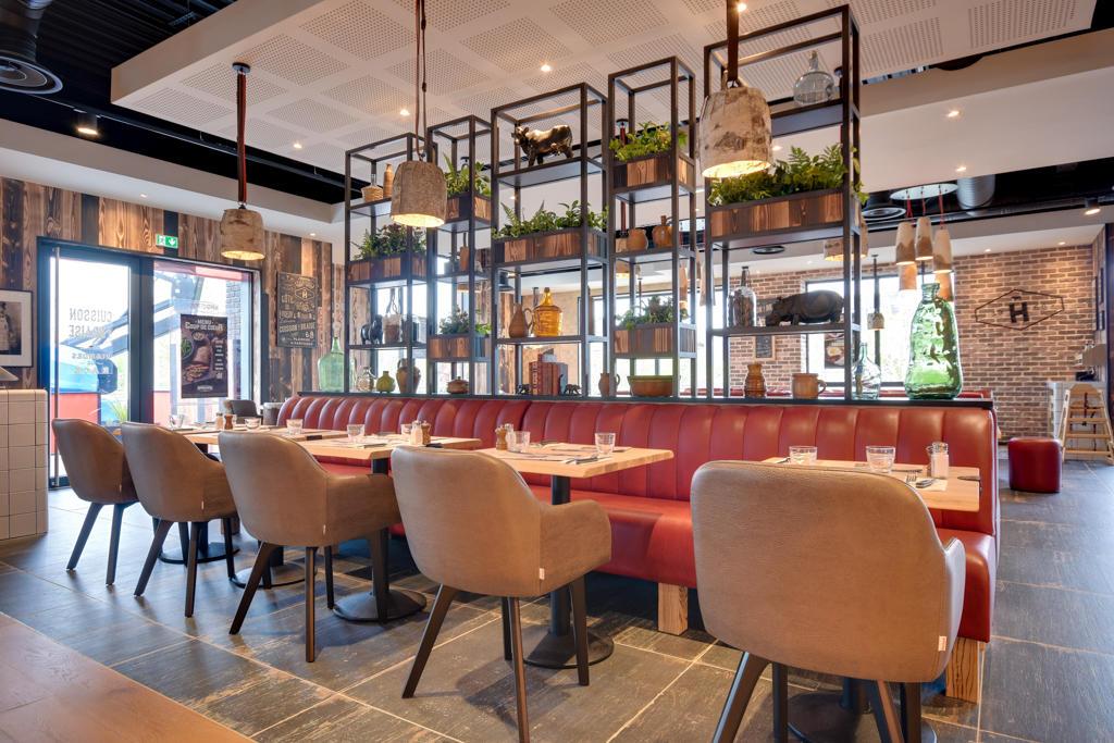 Un restaurant Hippopotamus moderne avec des sièges rembourrés rouges, des chaises marron et des étagères ornées de plantes et d'objets décoratifs. Des tons ocres et un éclairage élégant.