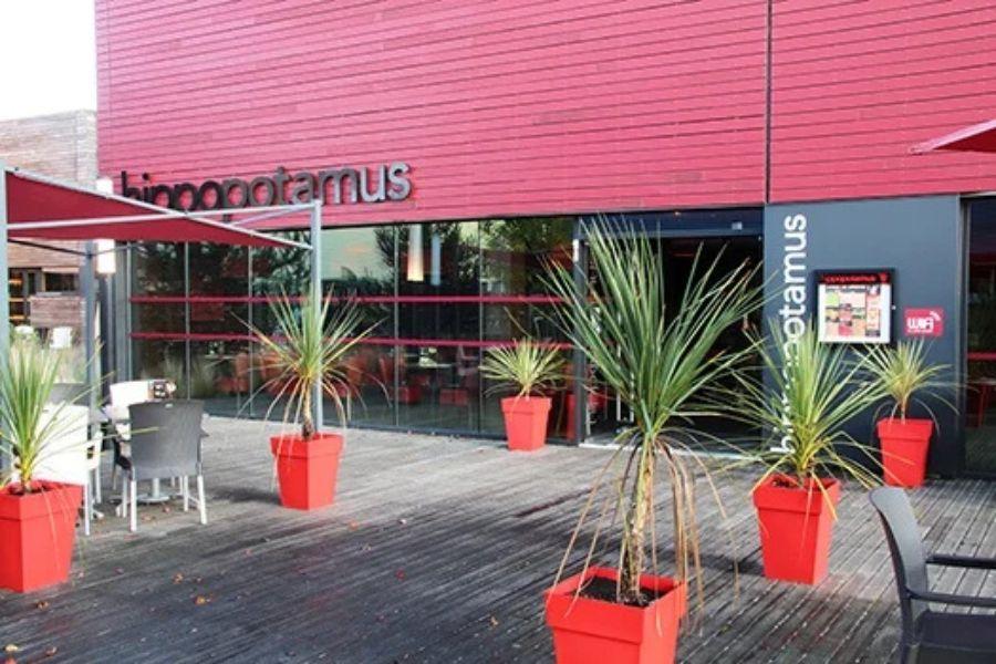 Extérieur du restaurant Hippopotamus avec des panneaux rouges et des jardinières rouges, agrémenté de grandes plantes vertes. Terrasse avec tables et chaises sur une terrasse en bois.