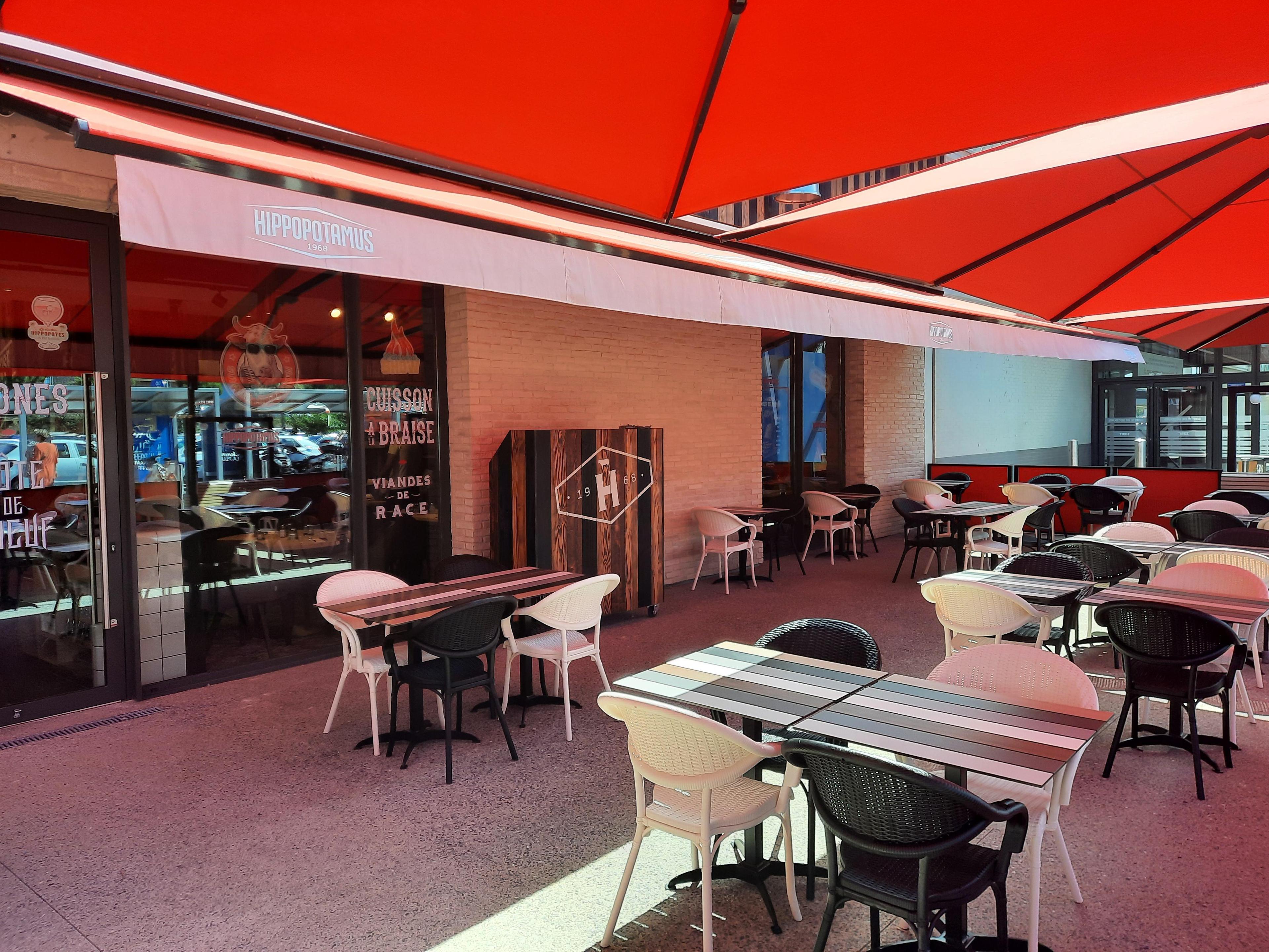 Restaurant Hippopotamus avec une terrasse extérieur avec des tables rayées et un mélange de chaises noires et blanches sous des parasols rouges. Vitrine en verre et décoration intérieure visibles à l'arrière-plan.