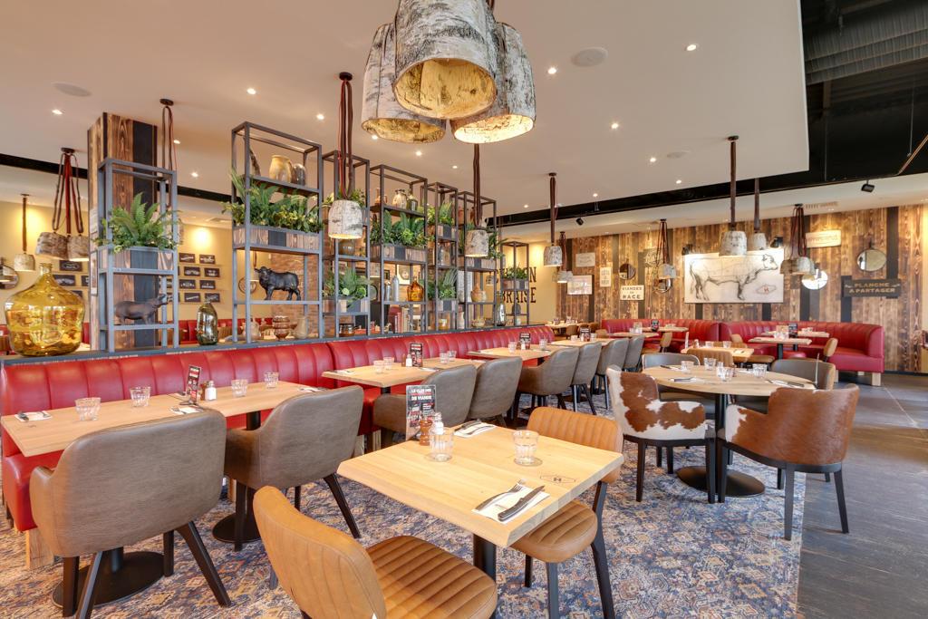 Intérieur de restaurant Hippopotamus moderne avec banquettes rouges, tables en bois, chaises assorties, suspensions lumineuses et étagères décoratives pour plantes. Touches rustiques partout.
