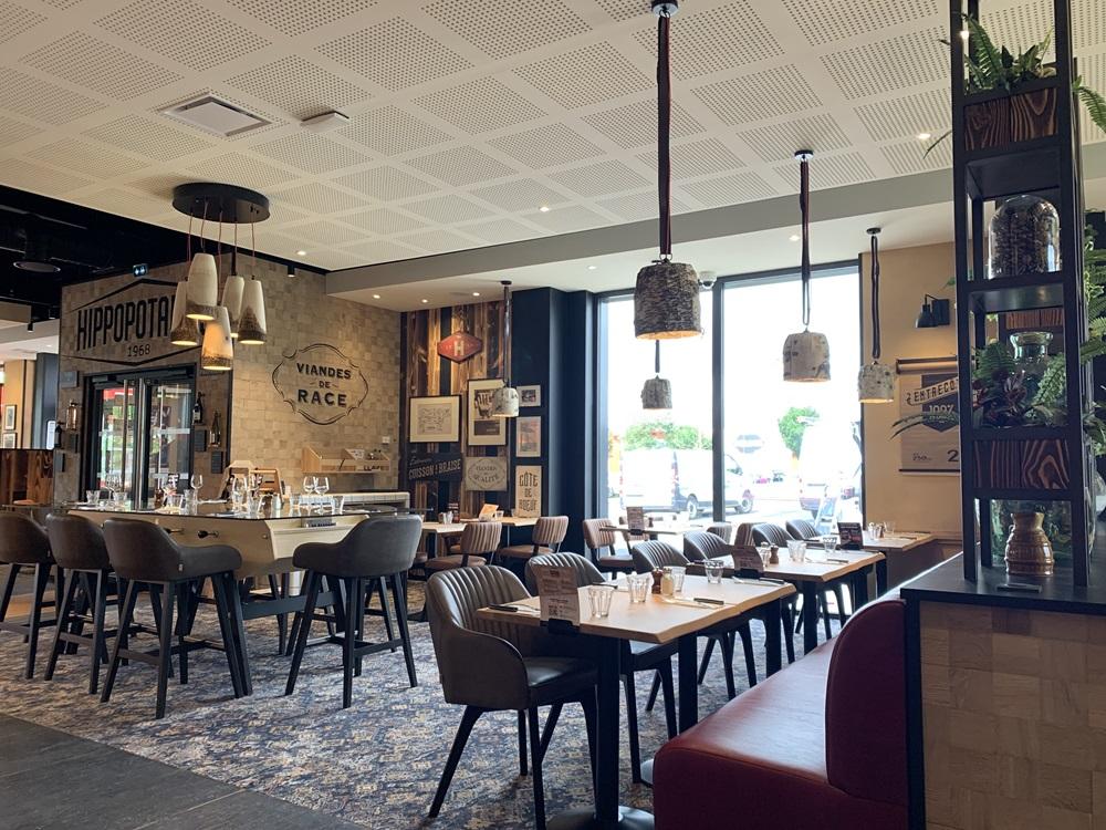 Intérieur moderne de restaurant Hippopotamus avec tables en bois, chaises rembourrées, suspensions lumineuses et grande fenêtre. Murs ornés d'œuvres d'art encadrées et de plantes vertes.