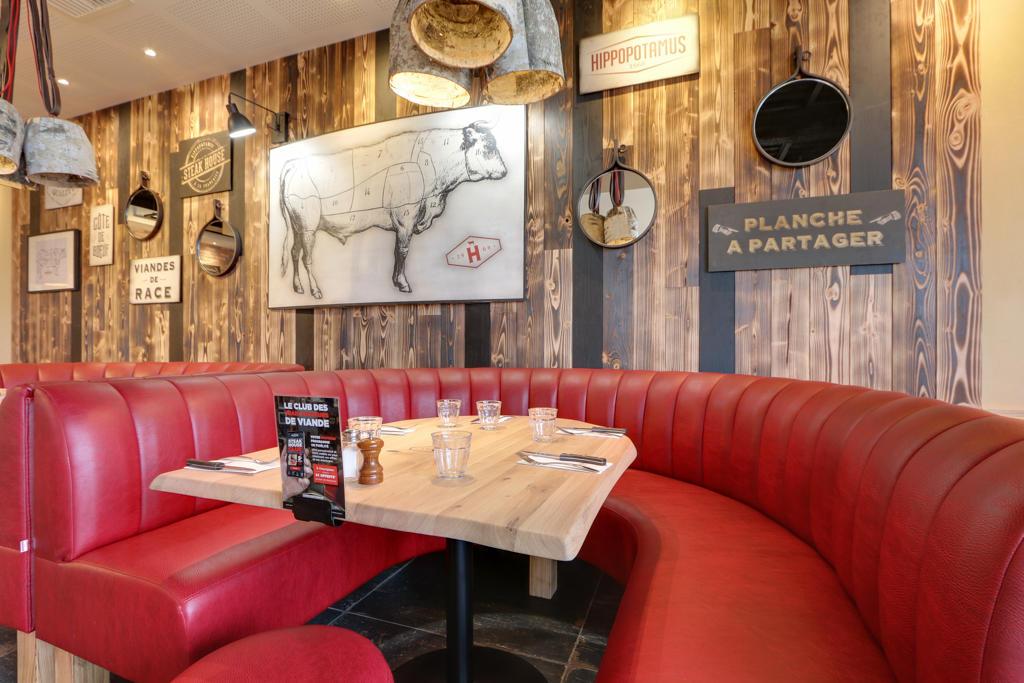 Intérieur de restaurant Hippopotamus moderne avec banquettes rouges, tables en bois, chaises assorties, suspensions lumineuses et étagères décoratives pour plantes. Touches rustiques partout.