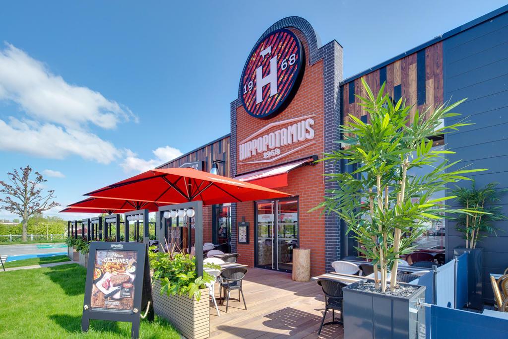 Extérieur d'un restaurant Hippopotamus avec des parasols rouges, des tables en terrasse et une végétation luxuriante. Logo bien visible au-dessus de l'entrée et ciel dégagé en arrière-plan.