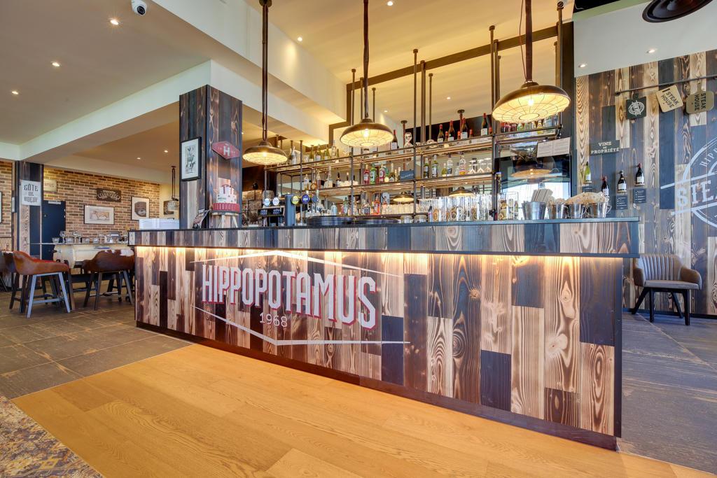 Restaurant Hippopotamus avec un intérieur de bar moderne avec un thème bois. Il comprend des suspensions lumineuses, des étagères avec des bouteilles et un coin salon confortable.