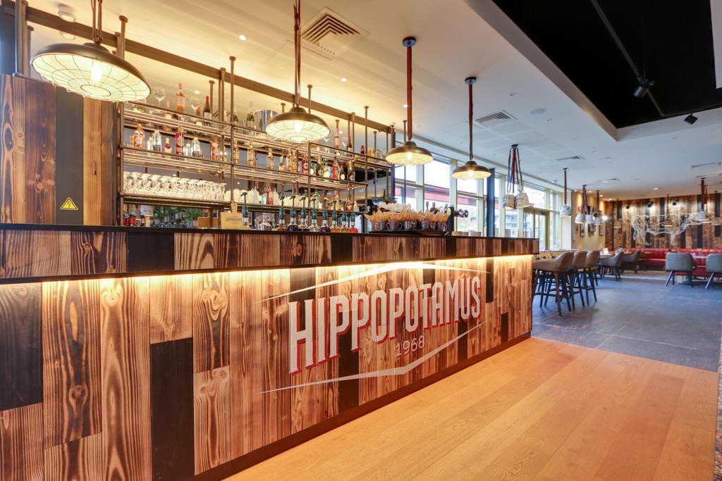 Intérieur moderne d'un restaurant Hippopotamus avec un comptoir en bois, des étagères remplies de bouteilles, un éclairage chaleureux et un coin salon avec des tables.