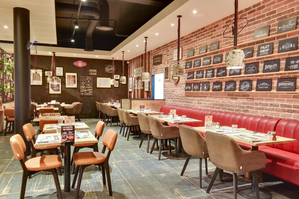 Intérieur de restaurant Hippopotamus chaleureux avec murs en briques, tables en bois, banquettes rembourrées rouges, menus écrits à la craie sur un tableau noir et éclairage tamisé.