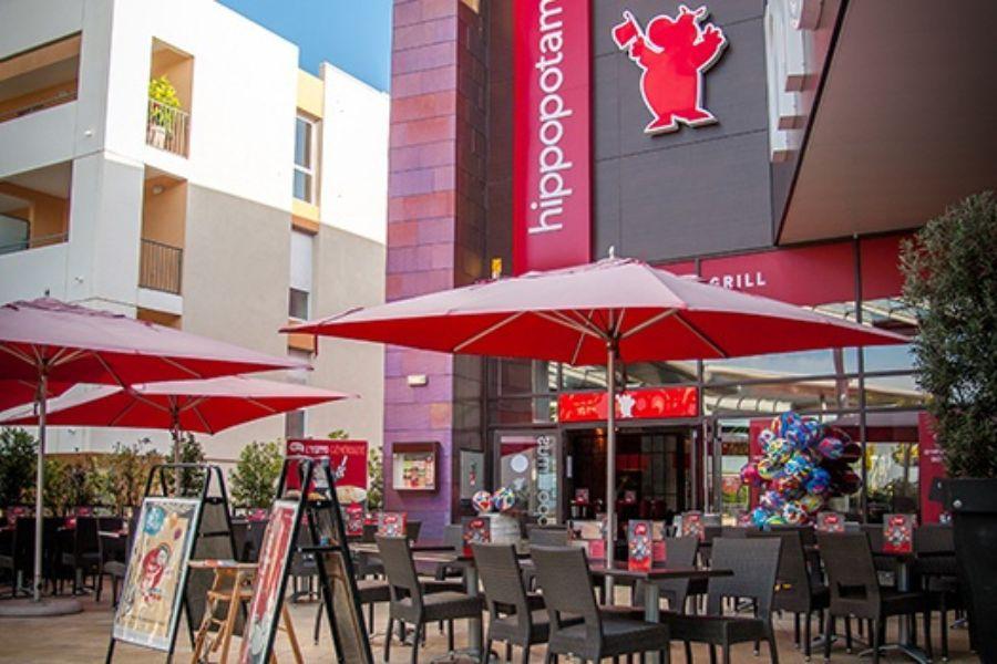 Terrasse extérieure du Hipopotamus avec parasols et chaises rouges, devanture avec logo rouge et ballons près de l'entrée.