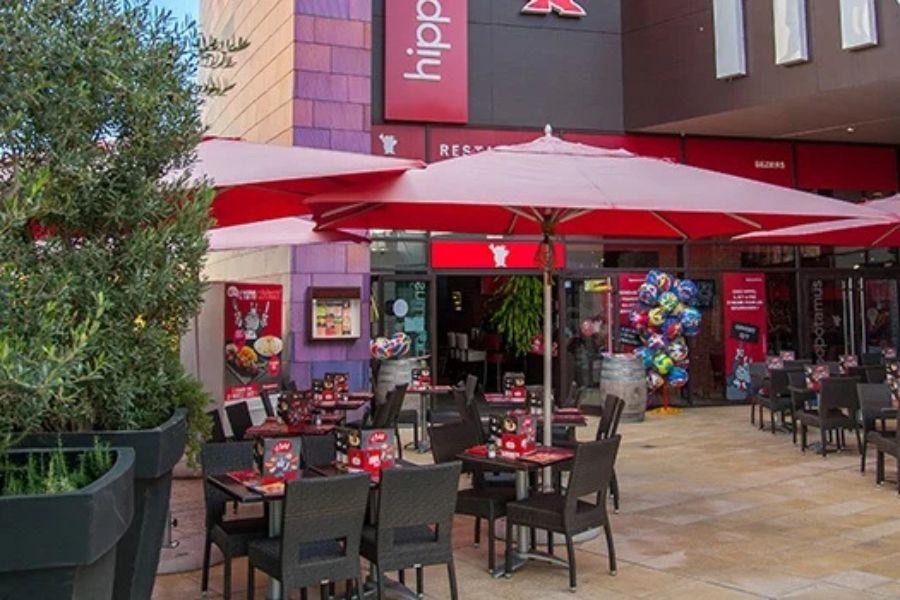 Espace repas extérieur avec parasols rouges, tables et chaises noires, adjacent à l'entrée d'un restaurant avec enseigne colorée et ballons.