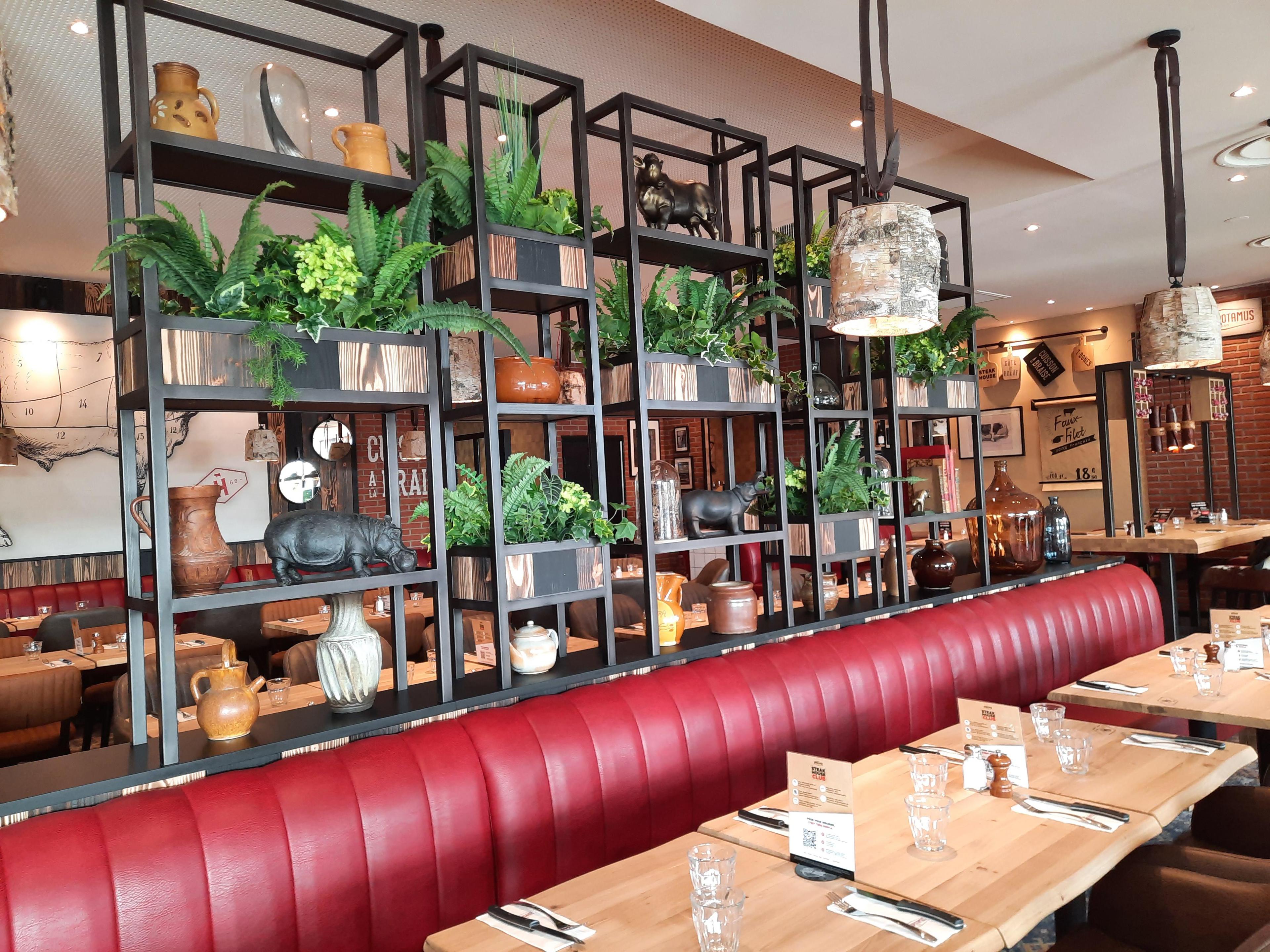 Intérieur de restaurant Hippopotamus avec sièges en cuir rouge, tables en bois et étagère décorative avec plantes et céramiques. Des suspensions industrielles sont suspendues au-dessus.